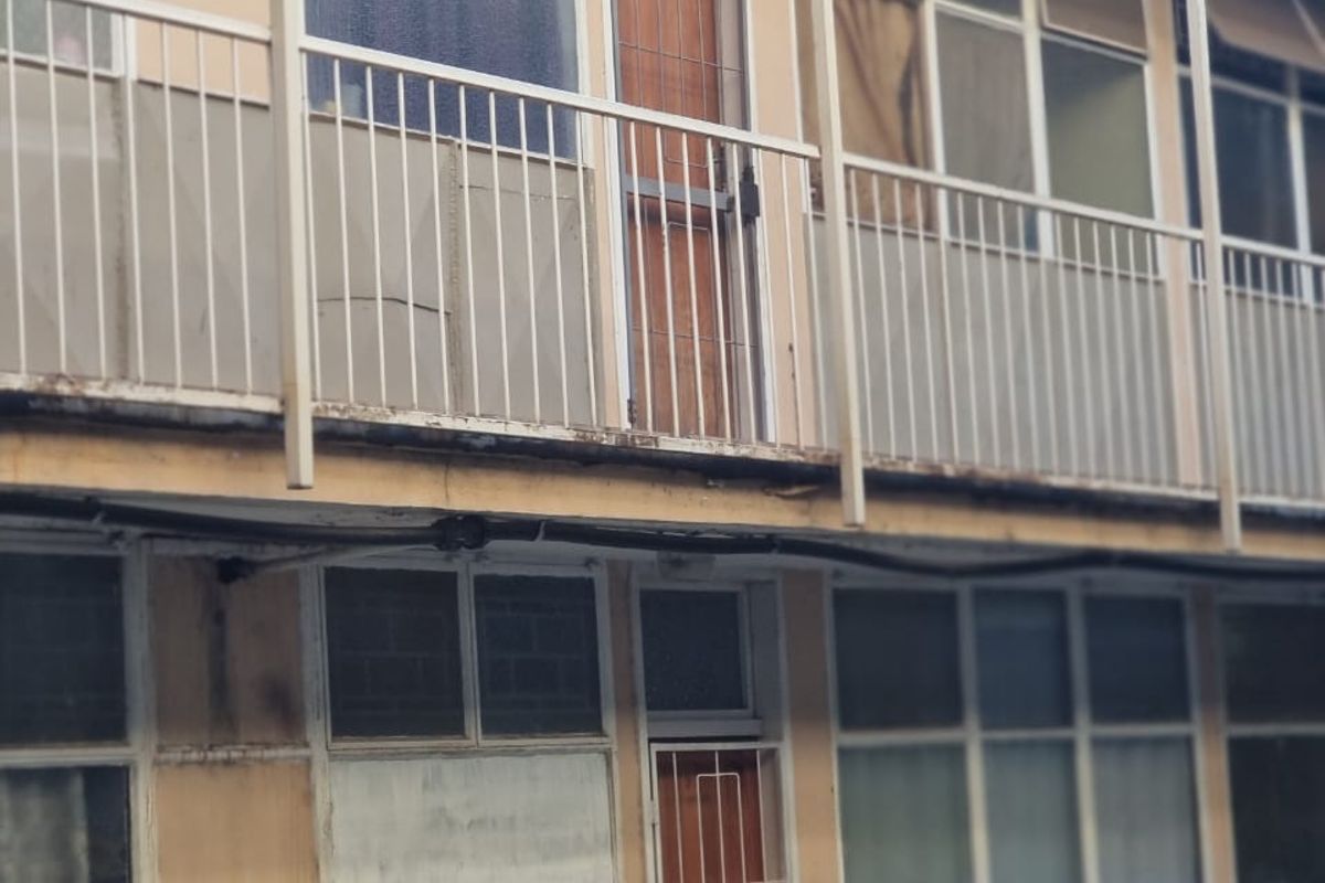 Exterior view of an apartment building facade featuring metal balcony railings, security gates, and multi-pane windows showing signs of significant weathering and wear.