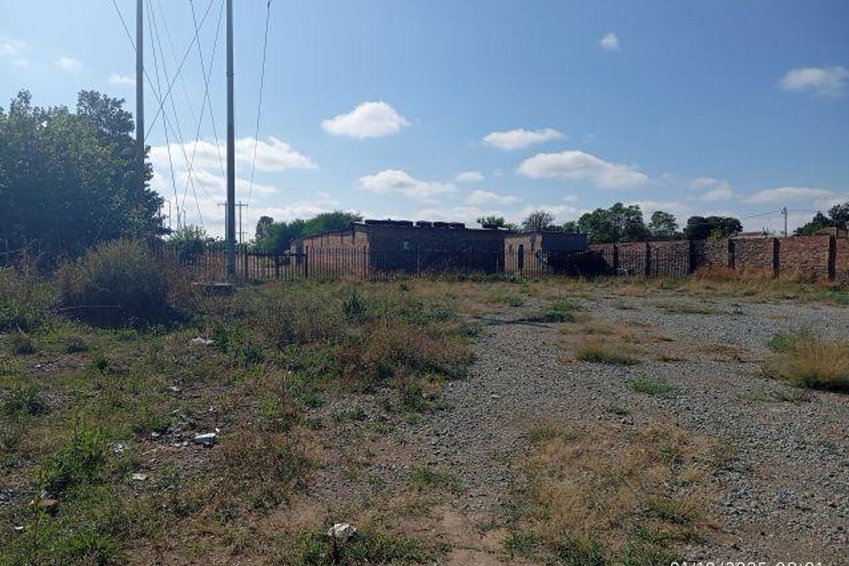 An expansive industrial plot featuring a brick perimeter wall, security fencing, and overhead power lines, currently characterized by unpaved ground and overgrown vegetation.