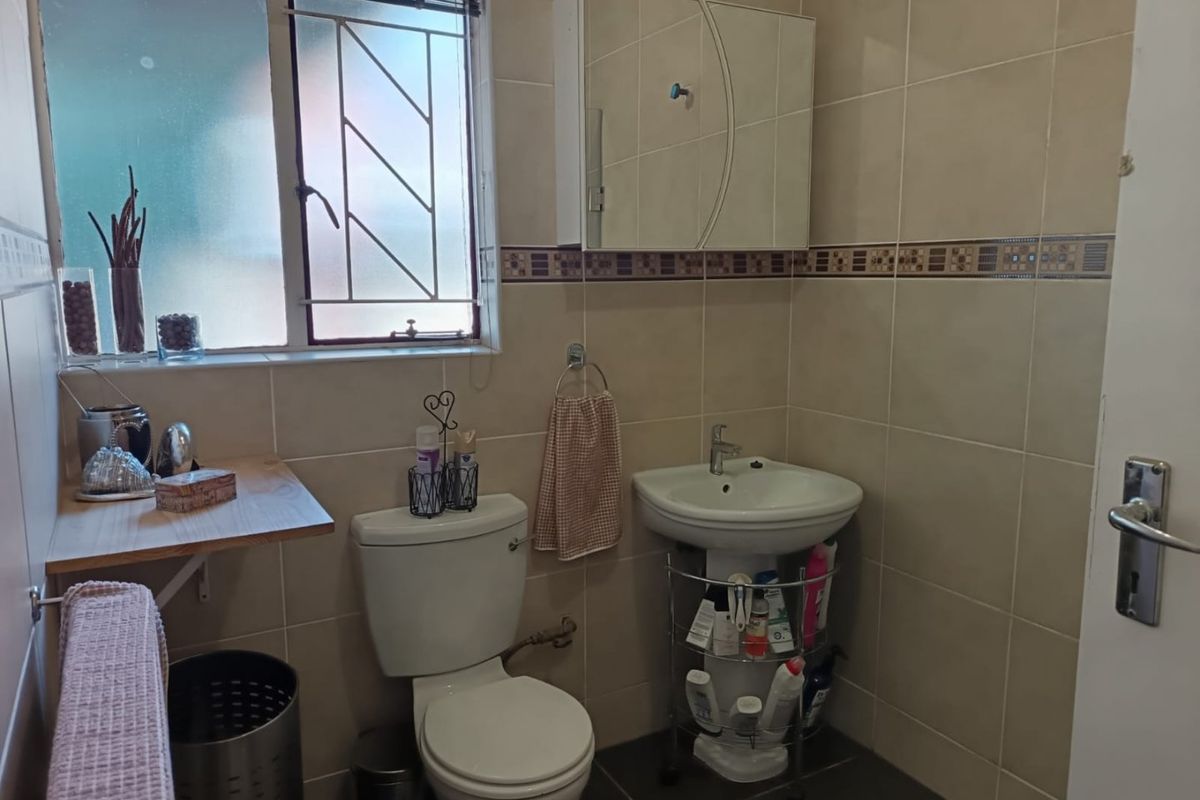 A functional bathroom featuring a pedestal basin, toilet, and wall-mounted medicine cabinet with neutral wall tiling and security bars on the window.