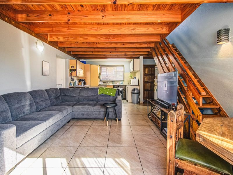 Open-plan living area featuring exposed timber ceiling beams, tiled flooring, and an integrated kitchen space with a timber staircase leading to the upper level.
