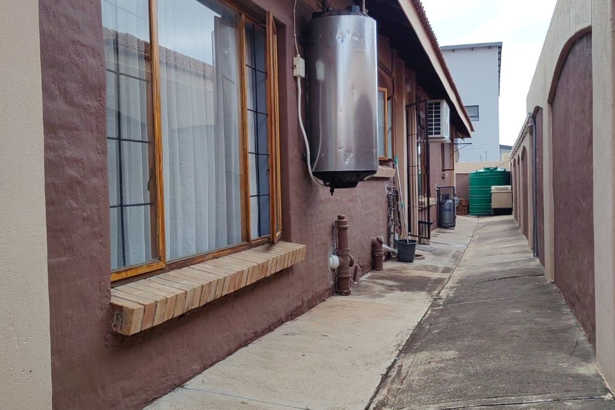 Side exterior view of the property featuring a concrete walkway, external geyser, and timber-framed windows along the building facade.