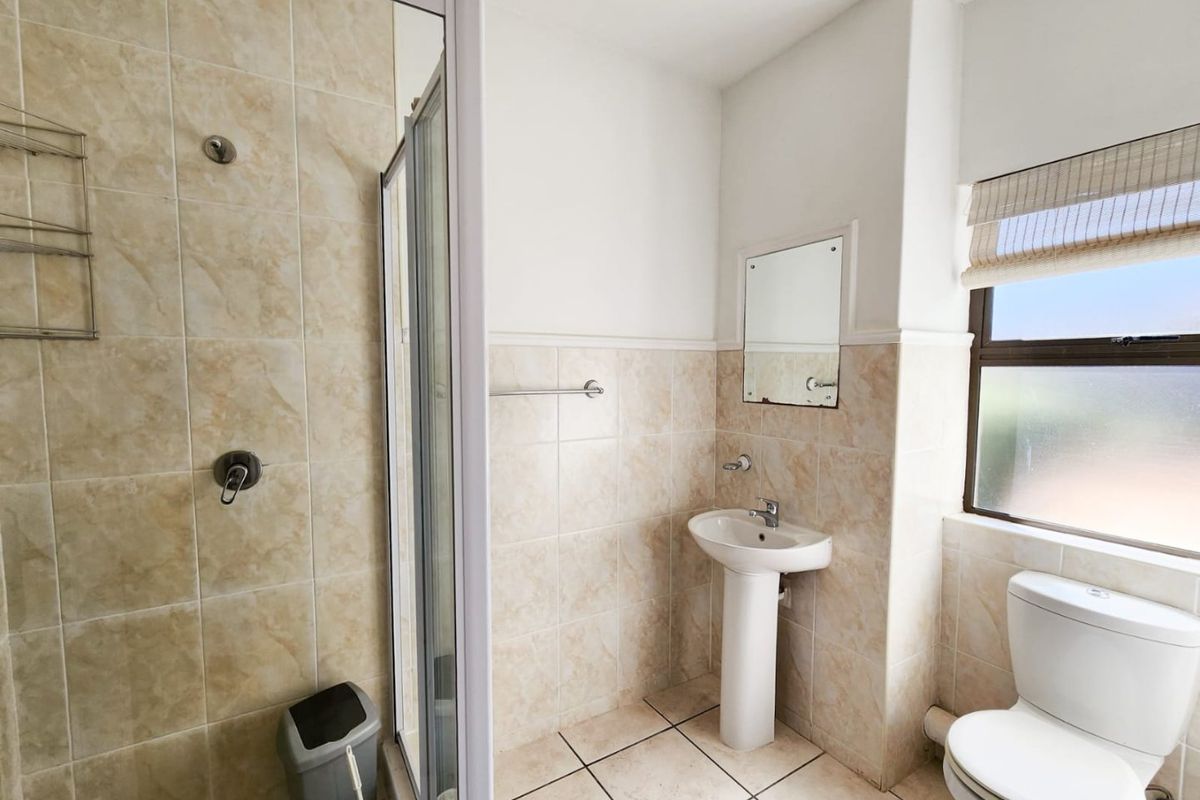 A functional bathroom featuring a pedestal basin, a glass-enclosed shower, and a toilet, finished with neutral wall and floor tiling.