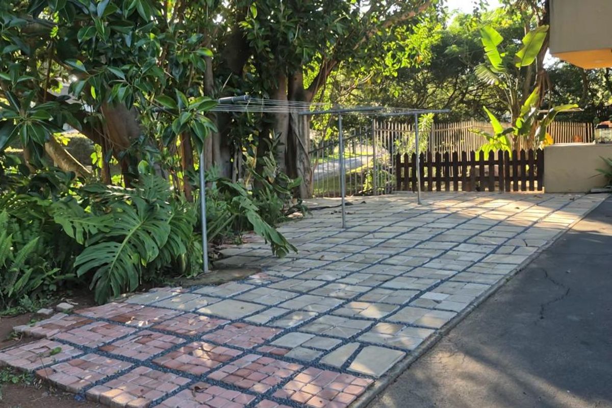 A rear outdoor area featuring a paved patio section, metal and wooden fencing, and mature garden trees.