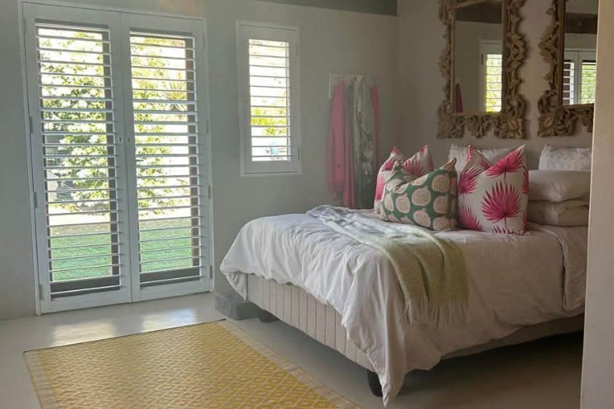 A bright bedroom featuring exposed timber ceiling beams, polished concrete flooring, and plantation shutters with direct access to the garden through french doors.