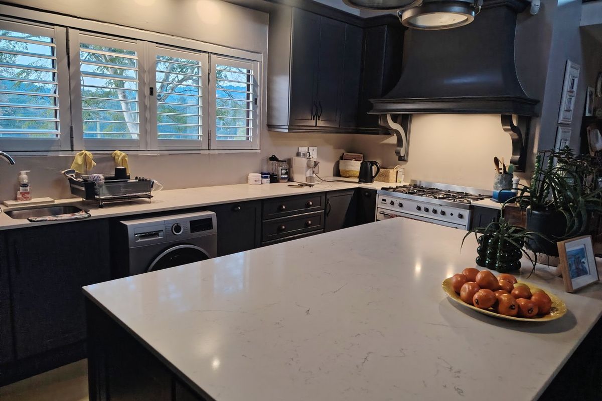 Modern kitchen featuring dark shaker cabinetry, white quartz countertops, a large central island, gas range with extractor hood, and plantation shutters.