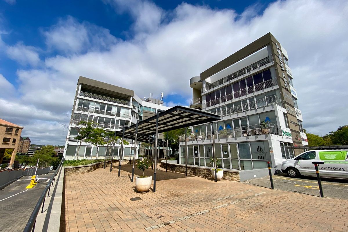 Modern multi-story commercial office building featuring a paved entrance courtyard with a covered walkway and extensive glass facade elements.
