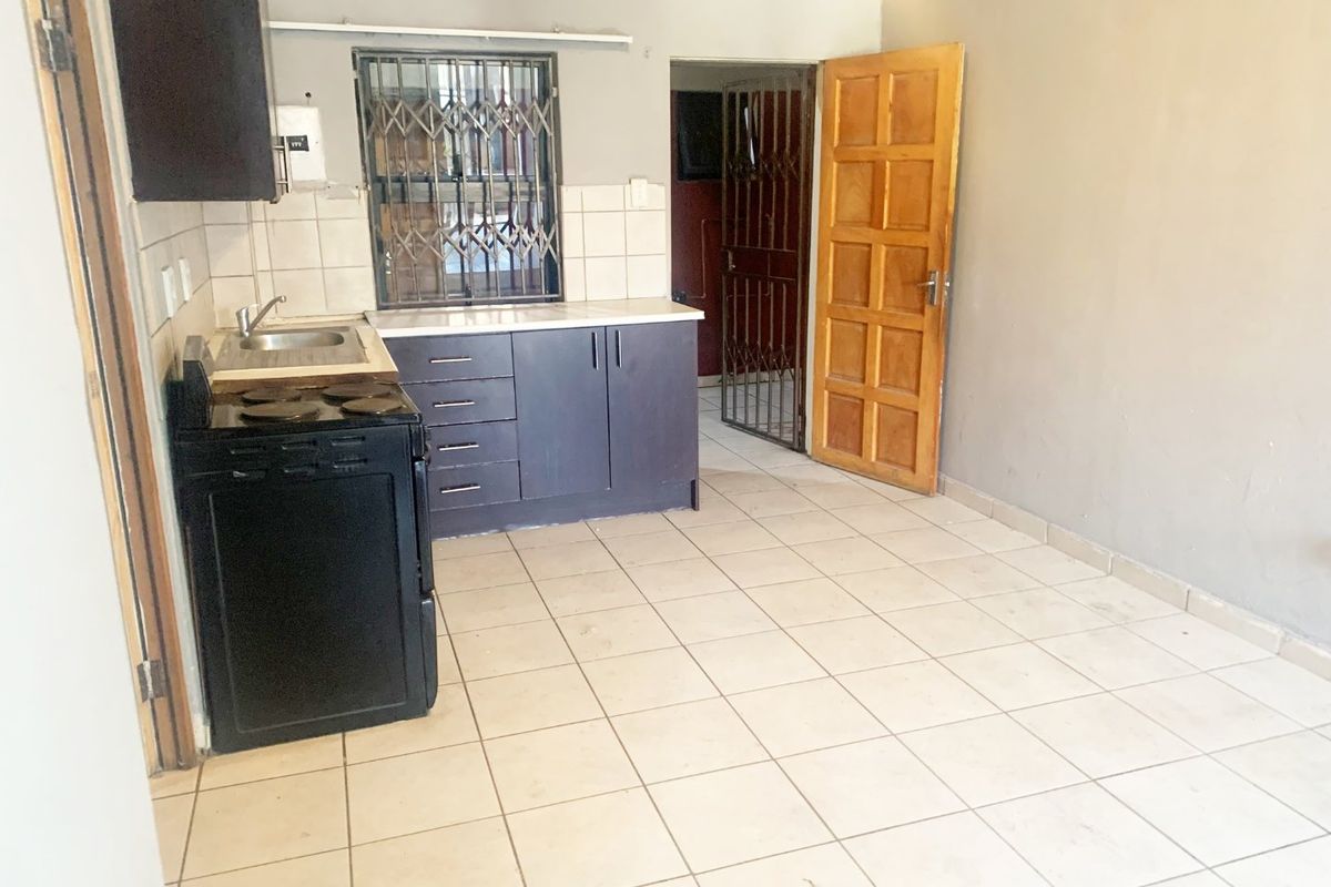 A compact kitchen area featuring tiled flooring, basic cabinetry, a stainless steel sink, and direct access to an adjacent room via a wooden door.