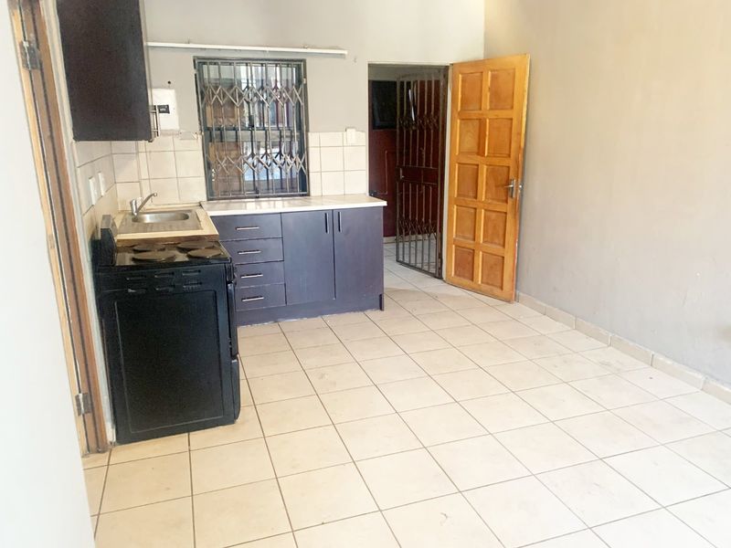A compact kitchen area featuring tiled flooring, basic cabinetry, a stainless steel sink, and direct access to an adjacent room via a wooden door.