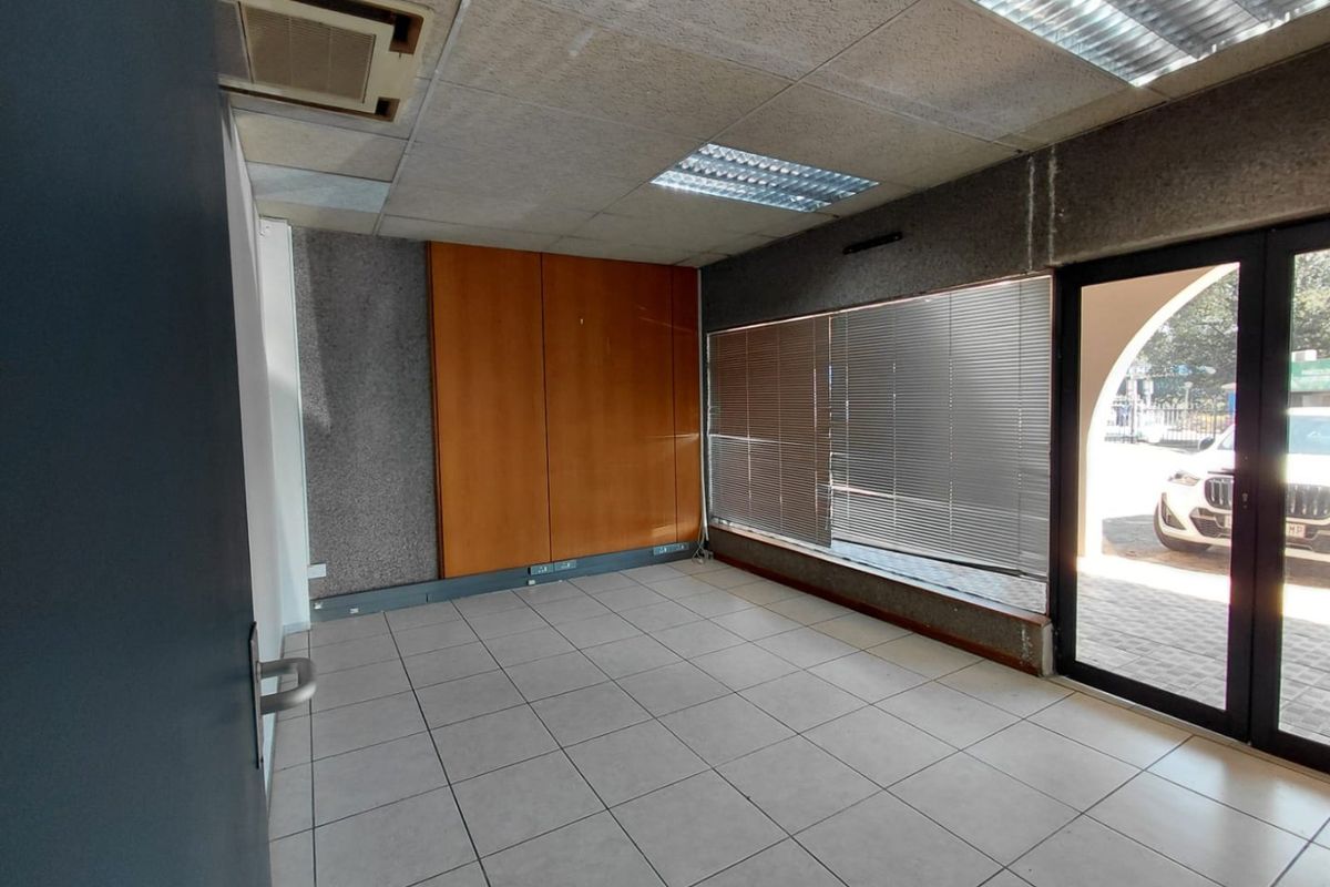 Empty commercial office space featuring tiled flooring, built-in wooden cabinetry, suspended ceiling with recessed lighting, and large windows with venetian blinds.