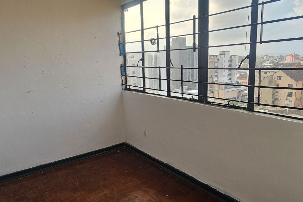 An empty bedroom featuring original parquet flooring, large casement windows with metal security bars, and urban city views.