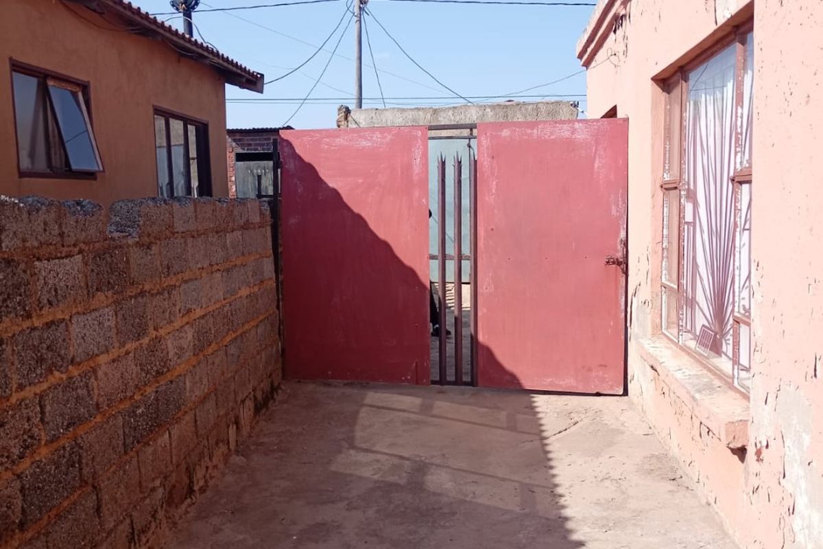 A narrow exterior side passage featuring a concrete boundary wall, metal gate, and weathered exterior walls with visible paint deterioration.