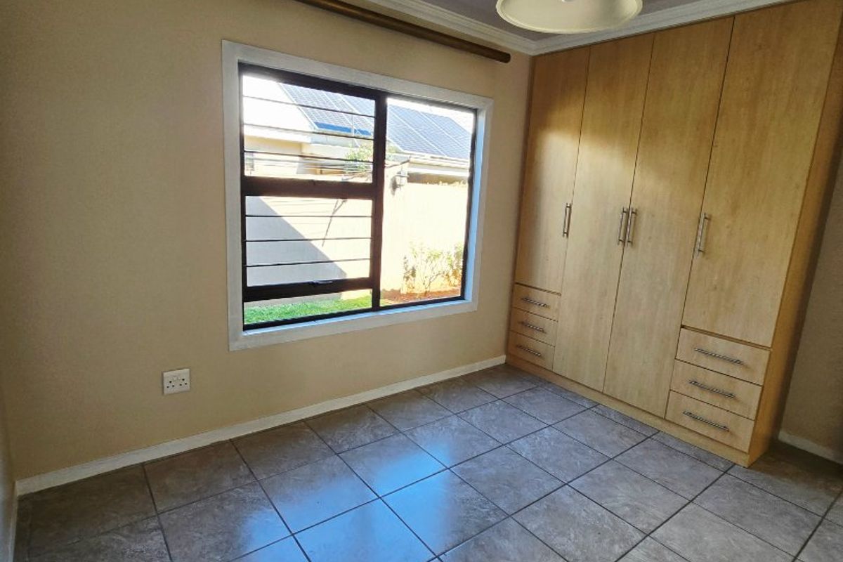 A clean, empty bedroom featuring neutral tiled flooring, built-in wooden cupboards, and a large window providing natural light.