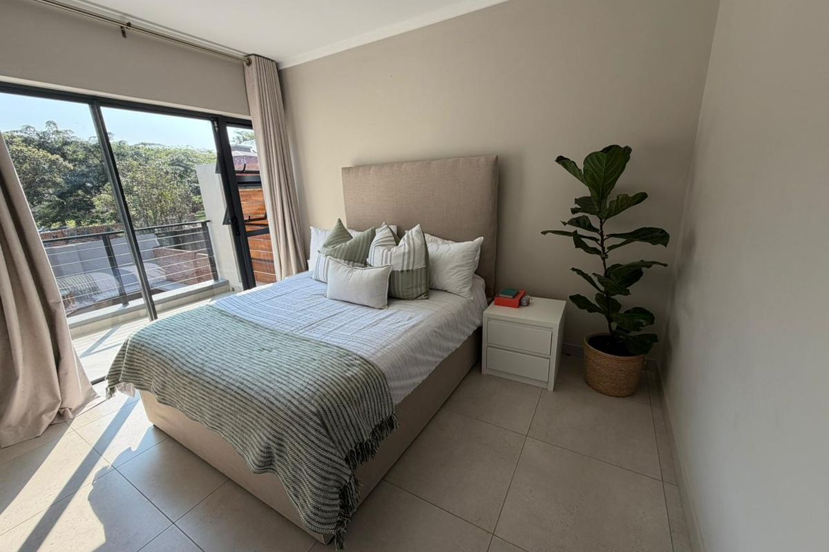 A bright bedroom featuring large sliding glass doors that open to a balcony, neutral tiled flooring, and a clean, modern aesthetic.