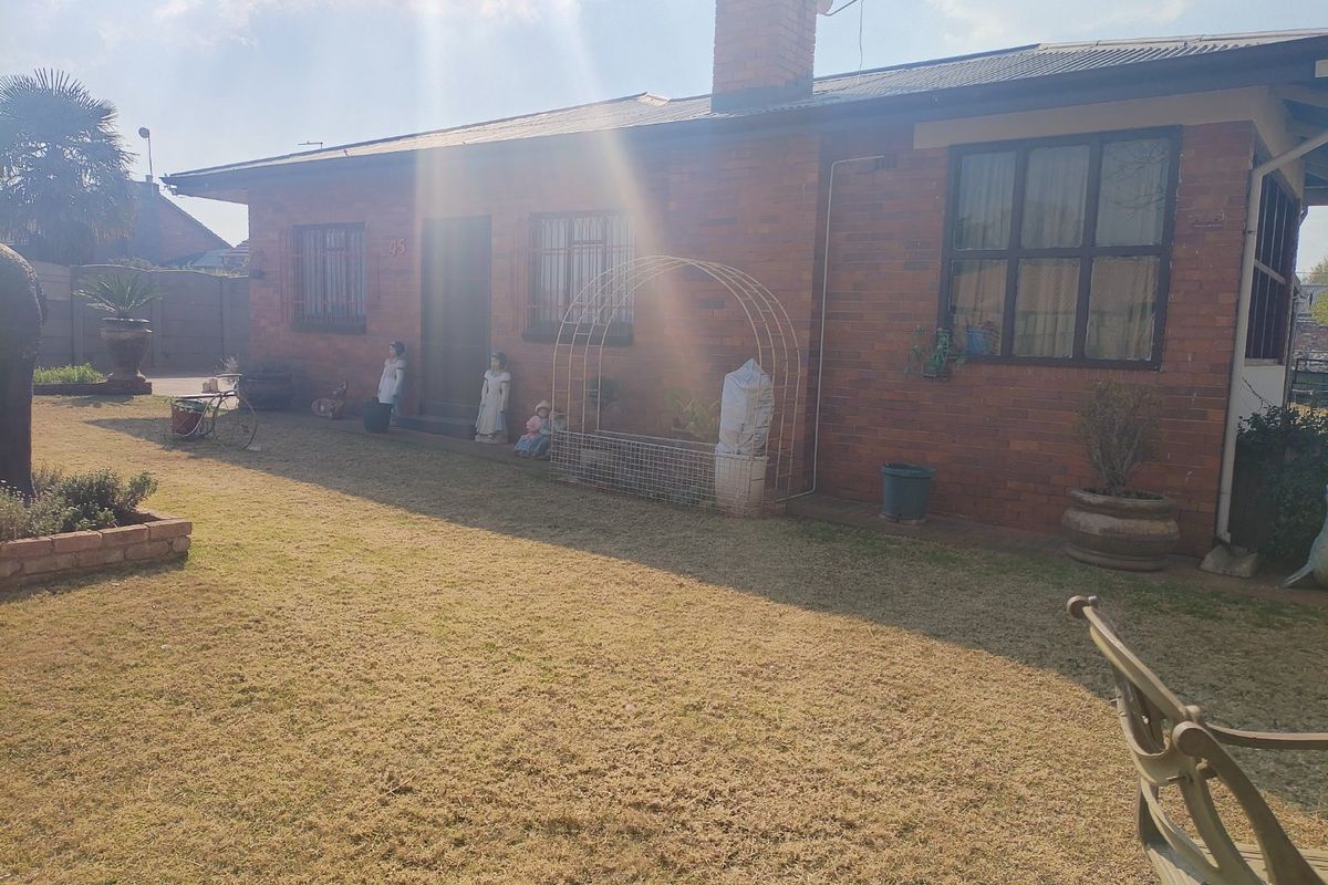 A rear view of a single-story brick house featuring a chimney, security bars on windows, and a surrounding garden area.