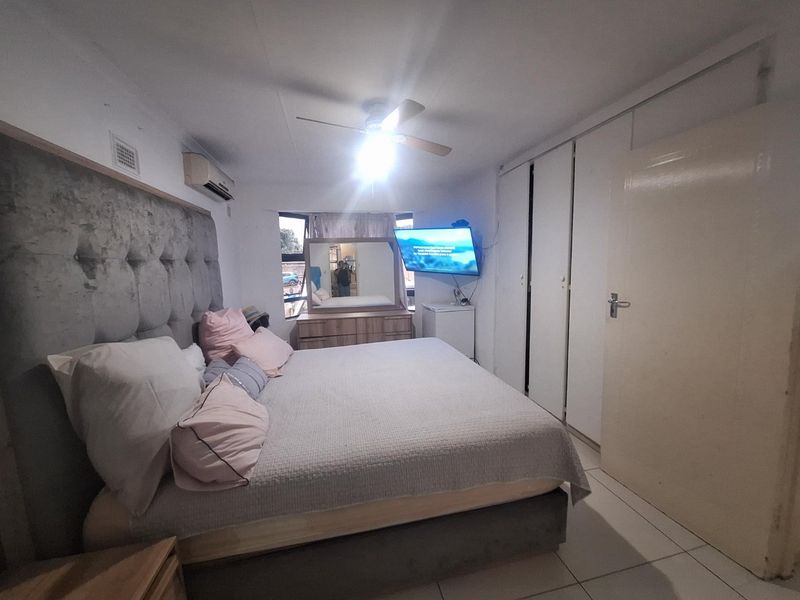 A functional bedroom featuring built-in cupboards, a ceiling fan, and an air conditioning unit, finished with tiled flooring.