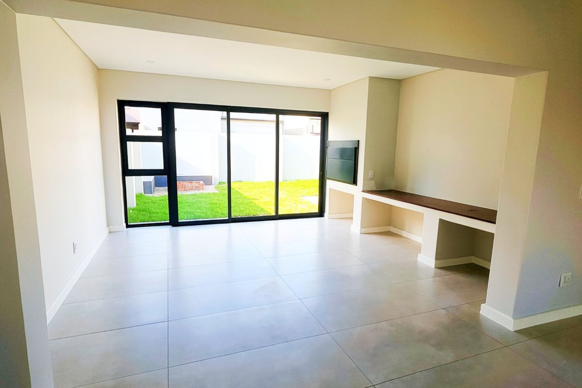 A bright, modern, and empty living room featuring large format floor tiles, a built-in media console, and sliding glass doors leading to the garden.