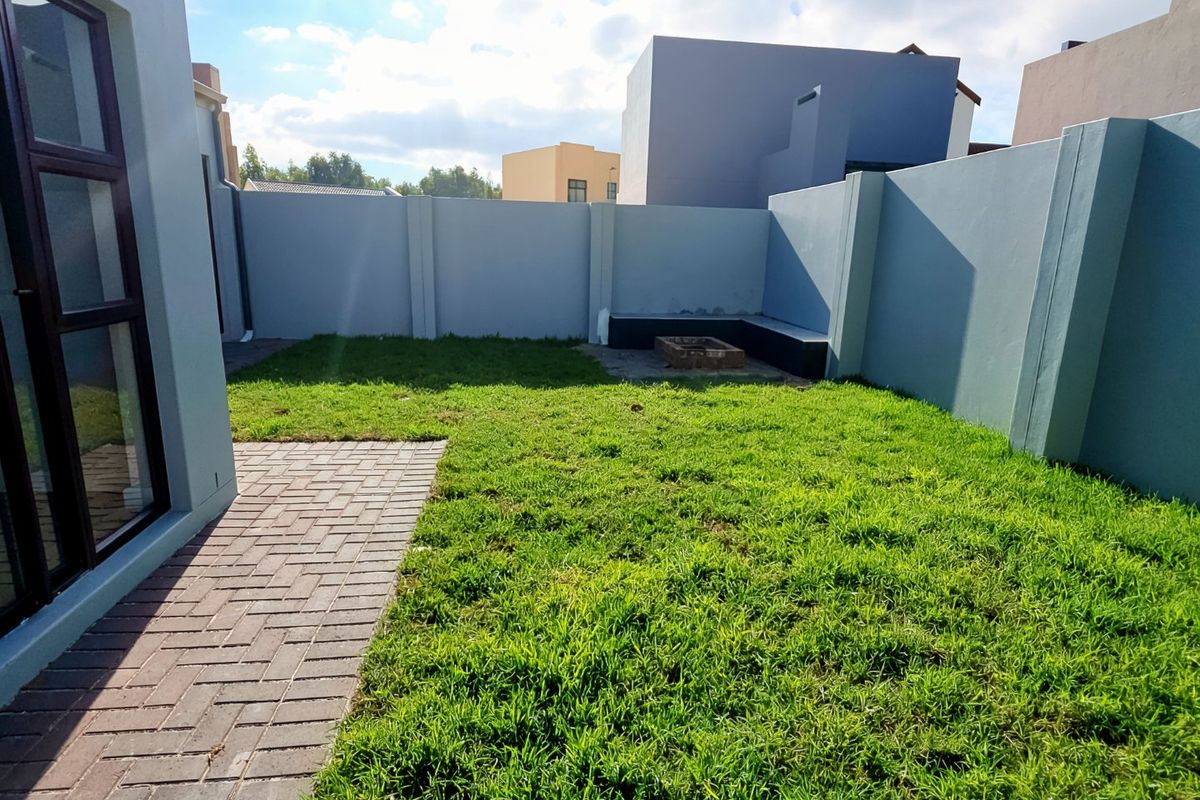 A private rear garden featuring a paved walkway, a grass lawn, and a built-in fire pit enclosed by high boundary walls.