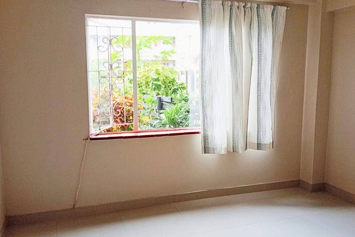 A bright, empty bedroom featuring tiled flooring, skirting boards, and a window with security bars overlooking greenery.