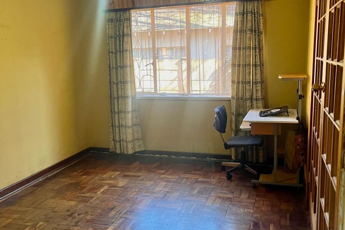 A bedroom featuring traditional parquet flooring, built-in wooden cupboards, and security bars on the window, offering a blank canvas for renovation.