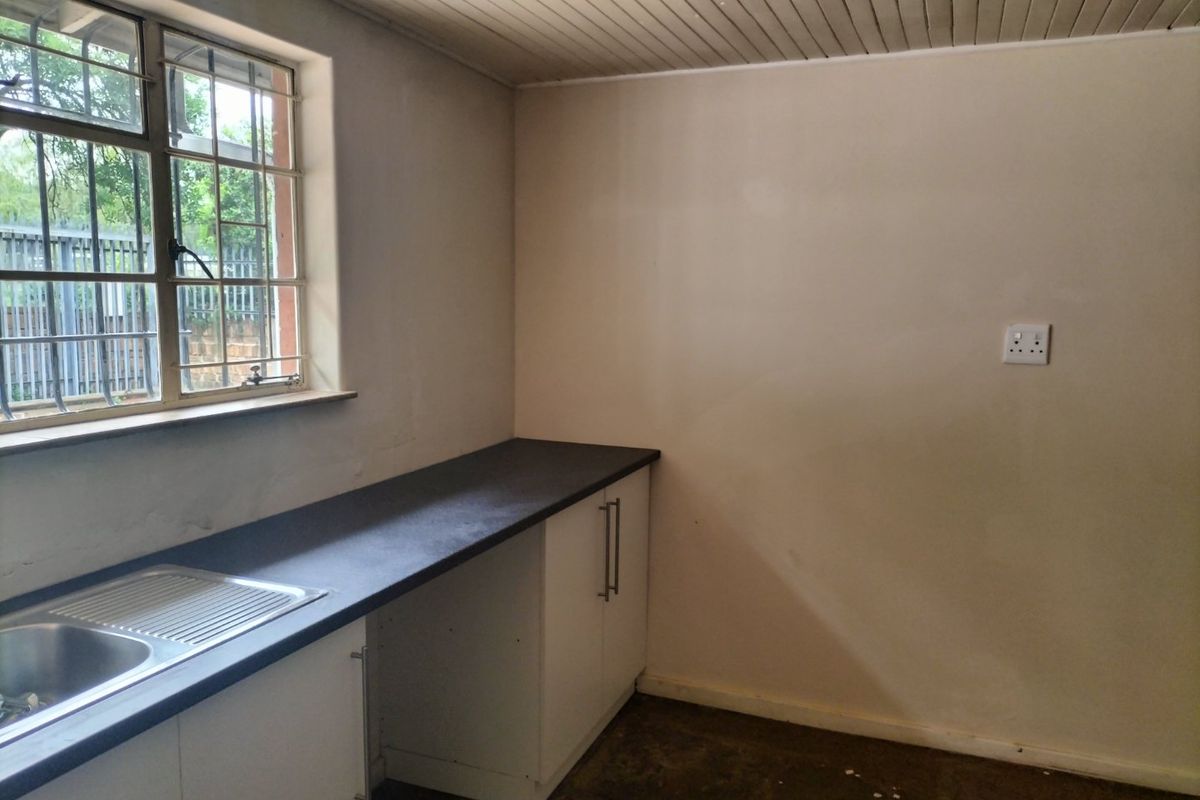 A compact, empty kitchen area featuring a stainless steel sink, dark laminate countertops, white base cabinetry, and a window with security bars.