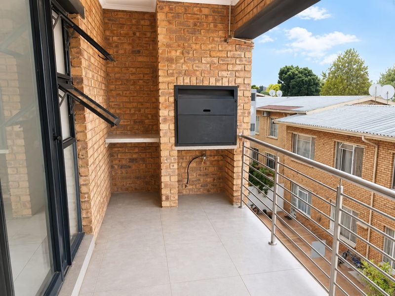 A covered balcony featuring a built-in braai, modern tiled flooring, and a stainless steel balustrade overlooking the apartment complex.