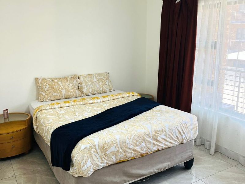 A well-lit bedroom featuring neutral tiled flooring and large windows with curtain rails, providing a clean and functional living space.