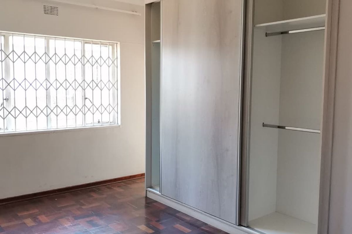 A vacant bedroom featuring parquet flooring, a built-in sliding door wardrobe, and a window fitted with security bars.