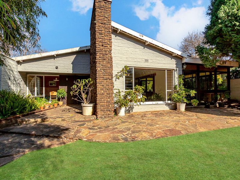 Rear view of the house featuring a stone patio, large picture window, and a prominent brick chimney set against a well-maintained garden.