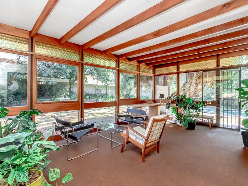 A bright, enclosed patio featuring exposed timber ceiling beams, extensive glass paneling, and direct access to the garden and pool area.
