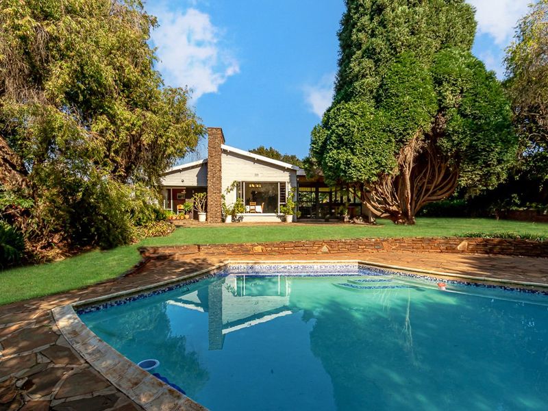 A well-maintained backyard featuring a swimming pool, stone patio, and a brick chimney facade set against a lush, mature garden.