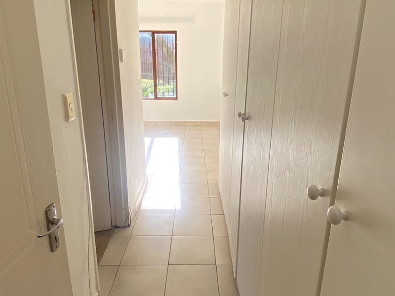 A bright, empty hallway featuring tiled flooring and extensive built-in cupboard storage, leading towards a sunlit room with security-barred windows.
