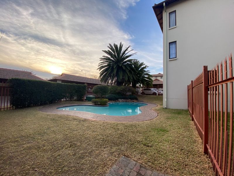 A well-maintained communal swimming pool area featuring brick coping, landscaped garden surrounds, and secure metal perimeter fencing within an apartment complex.