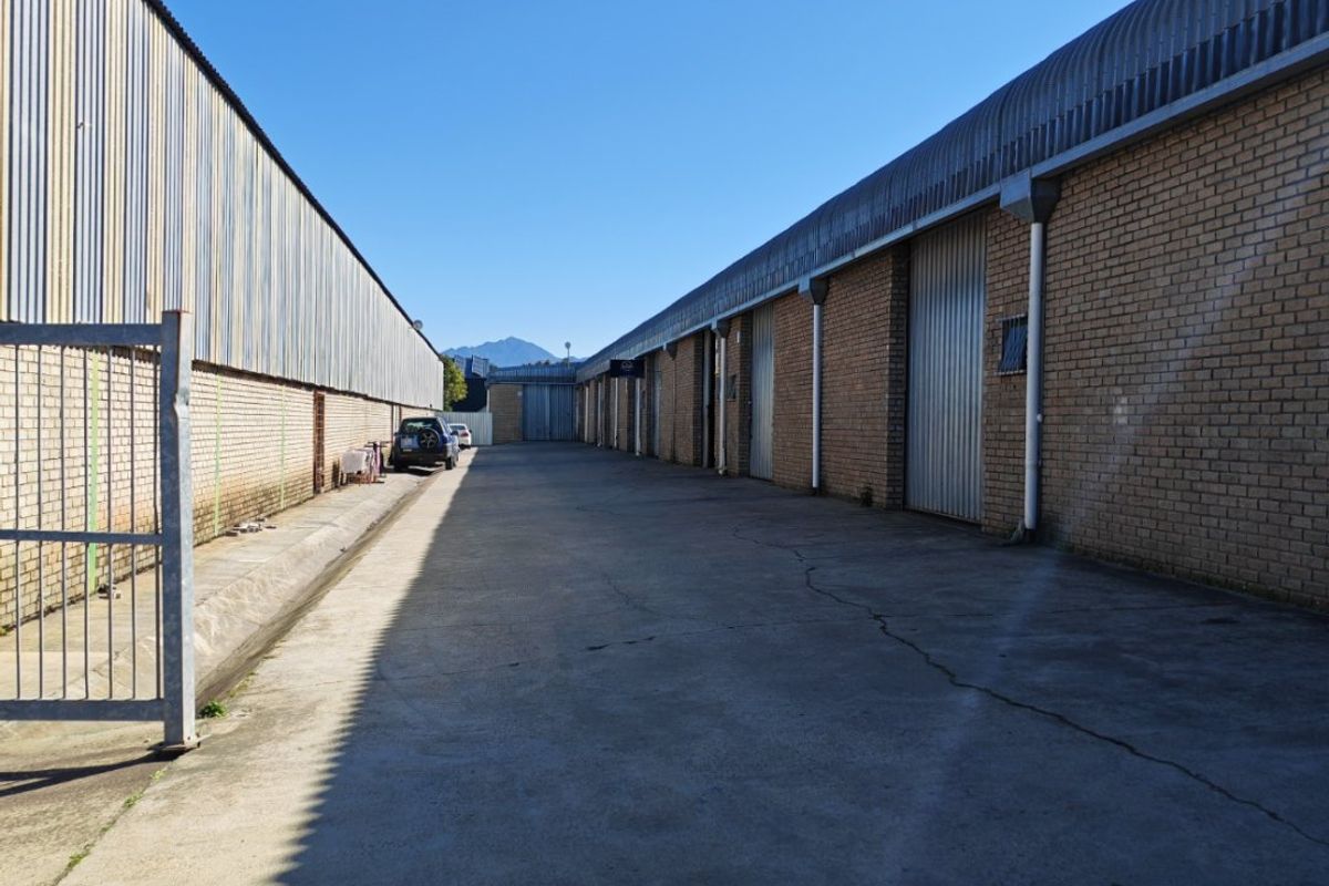 Industrial exterior view showing a concrete driveway between two warehouse buildings with brick walls and metal roller shutter doors.