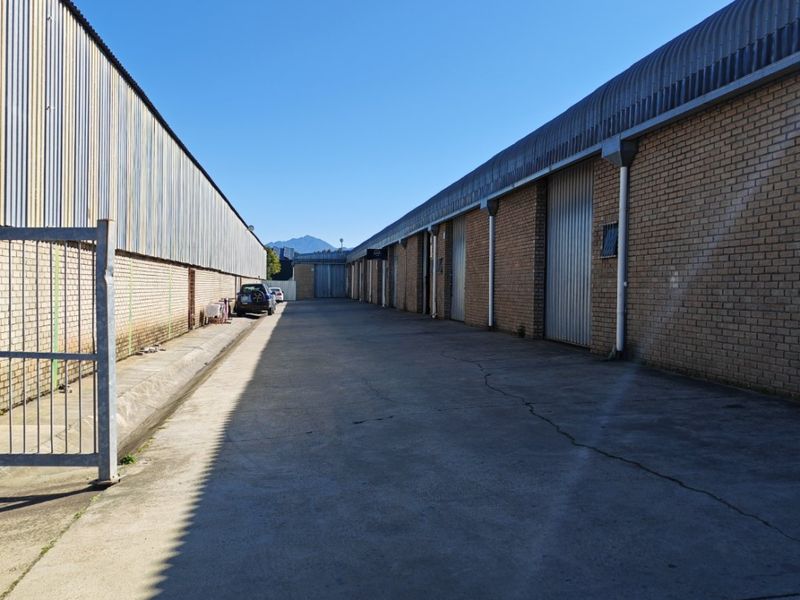 Industrial exterior view showing a concrete driveway between two warehouse buildings with brick walls and metal roller shutter doors.