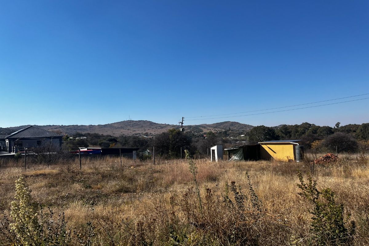 A view of a vacant residential plot featuring dry grass, wire fencing, and small utility structures against a mountain backdrop.