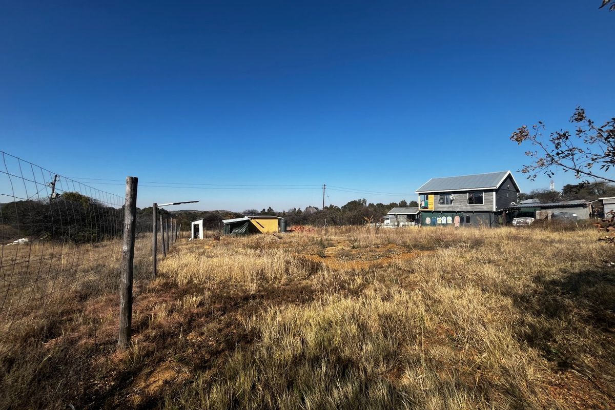 A wide view of a residential plot featuring dry grass, wire fencing, and existing outbuildings with a main dwelling in the background.