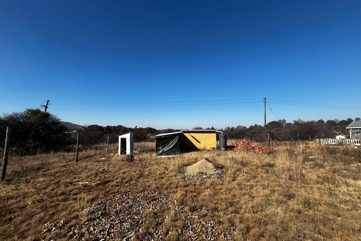A vacant residential plot featuring dry grass, a small utility shed, a water tank, and perimeter fencing under a clear blue sky.
