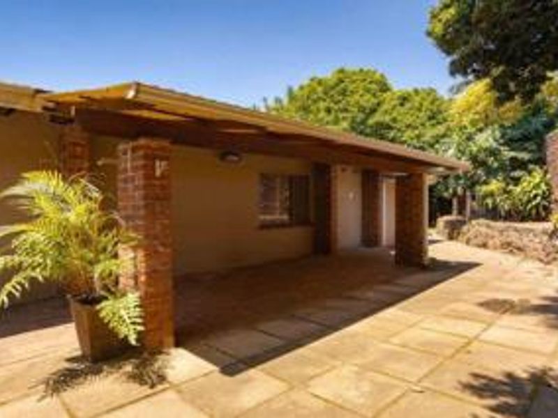 A covered outdoor patio area featuring brick pillars, timber pergola structure, and paved flooring, overlooking a landscaped garden with stone retaining walls.