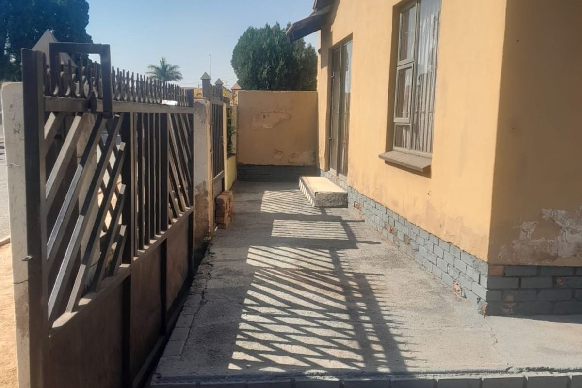 A side exterior view of the house featuring a metal security gate, concrete walkway, and visible wear to the exterior plaster and paintwork.