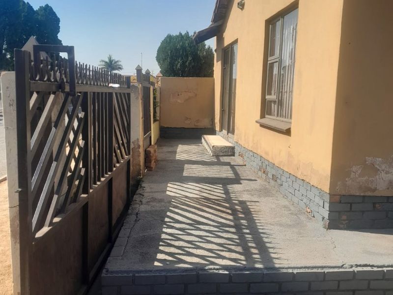 A side exterior view of the house featuring a metal security gate, concrete walkway, and visible wear to the exterior plaster and paintwork.