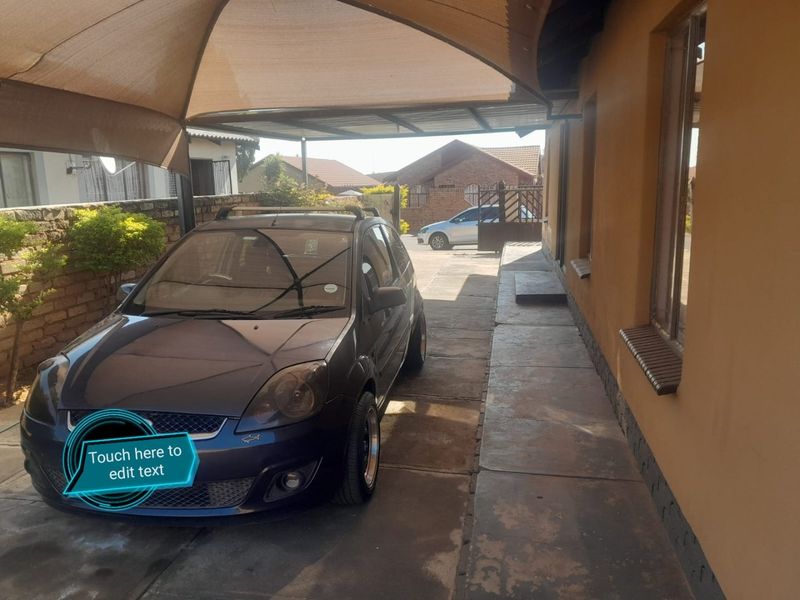A paved driveway area featuring a shade cloth carport structure and brick boundary walling, providing covered parking space.