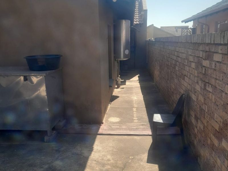 A narrow rear exterior walkway featuring a brick boundary wall, concrete paving, and an externally mounted electric water geyser.