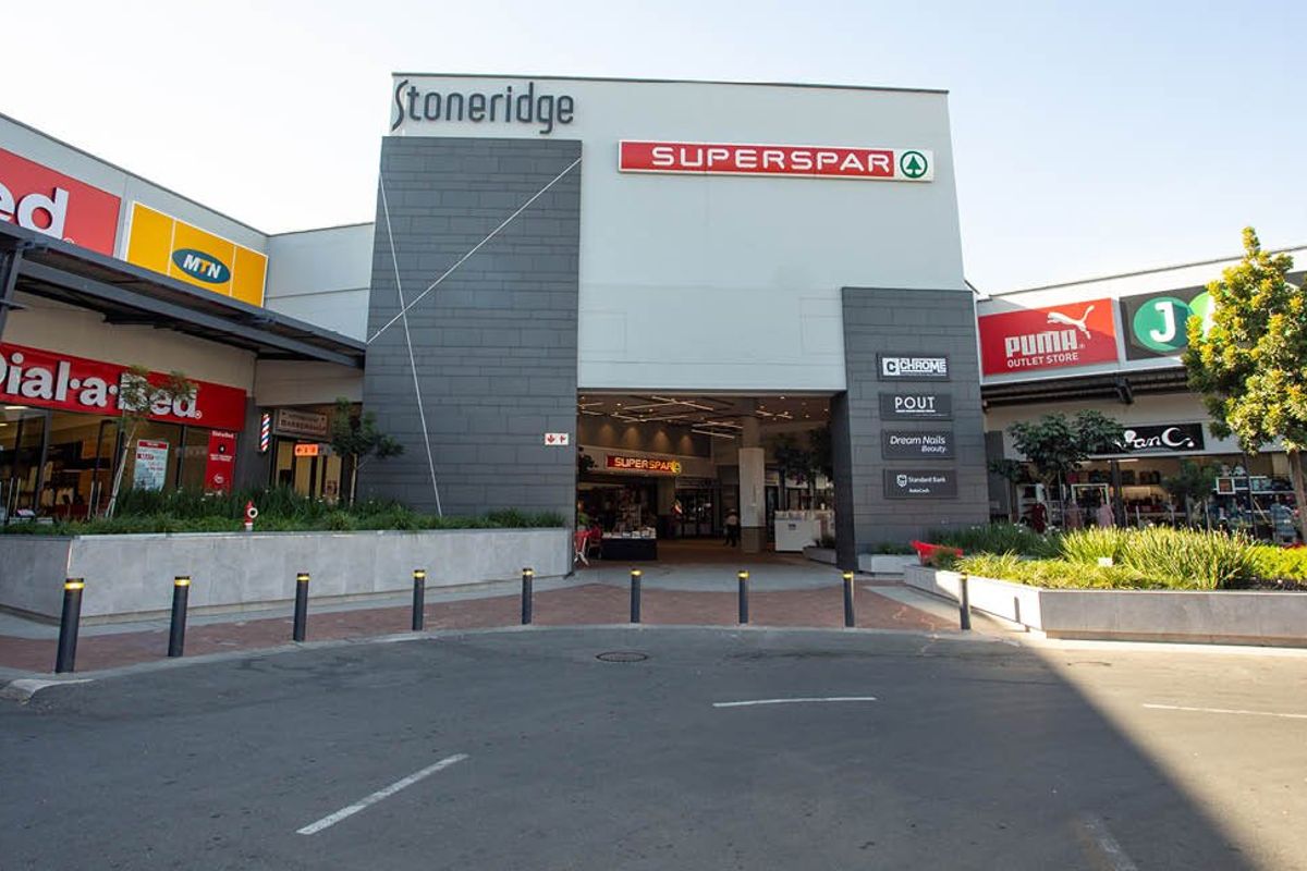 Front exterior view of Stoneridge commercial shopping center featuring multiple retail storefronts, prominent signage, and a paved entrance area with concrete bollards.