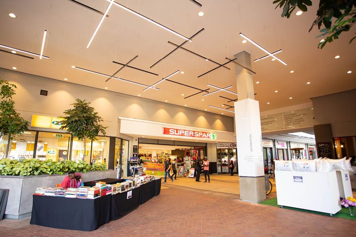 A modern commercial shopping center atrium featuring high ceilings with suspended linear lighting, brick flooring, and multiple retail storefronts.