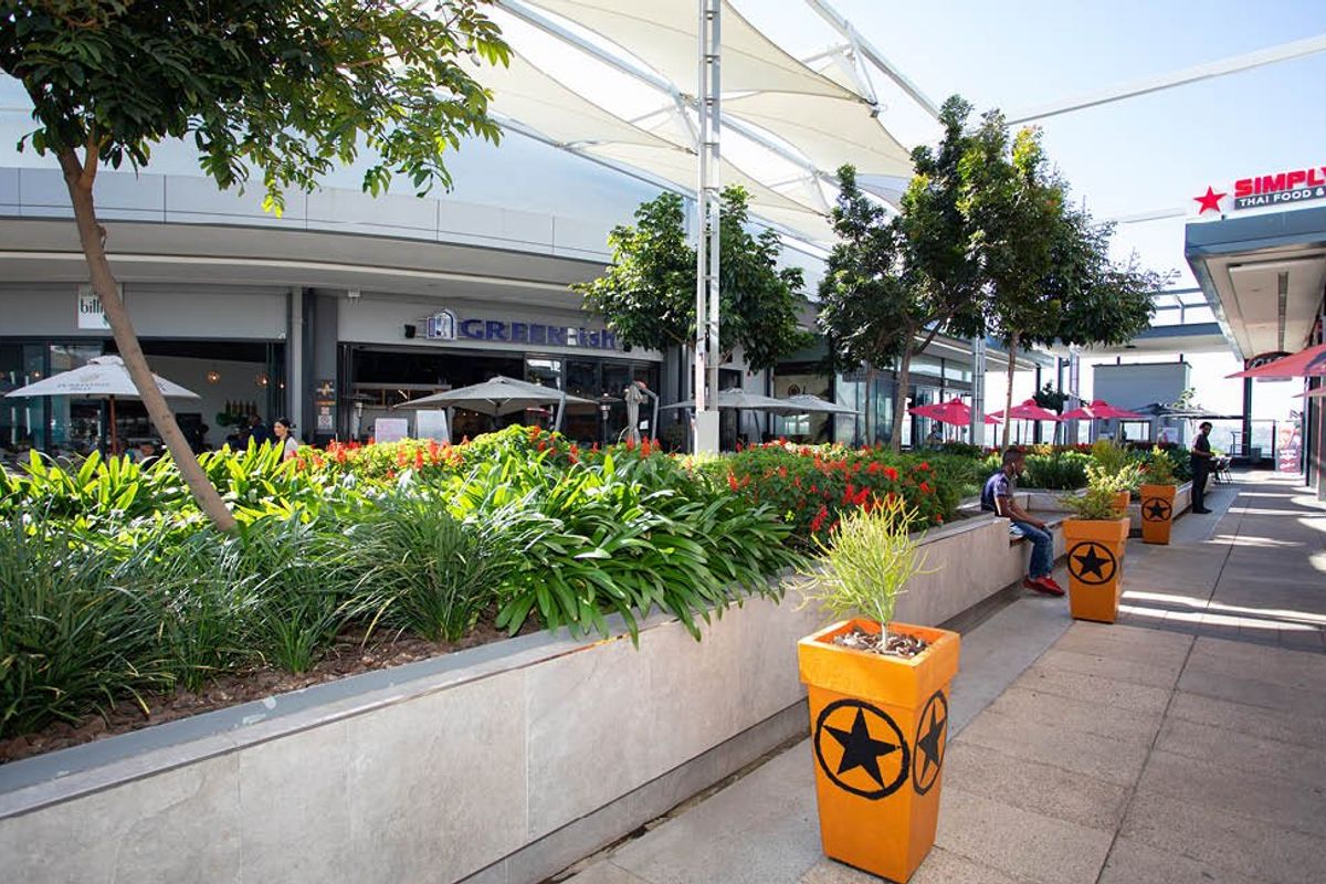 A well-maintained outdoor commercial promenade featuring tensile fabric shading, stone-clad planters, and modern storefronts in a vibrant retail environment.