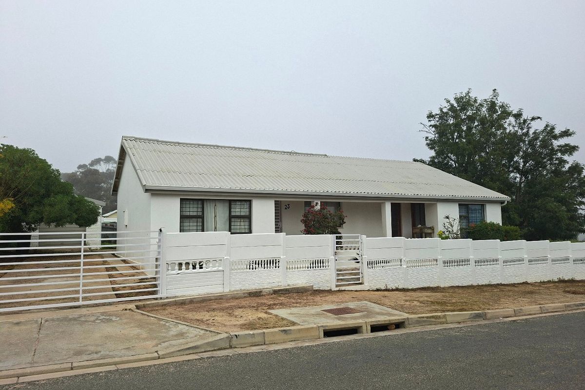 Front exterior view of a single-story house featuring a corrugated metal roof, white painted facade, and a secure precast concrete boundary wall with driveway gate.