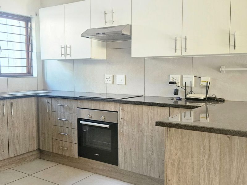 A functional kitchen featuring light wood-grain cabinetry, dark granite countertops, built-in oven, electric hob, and a stainless steel sink with tiled splashbacks.