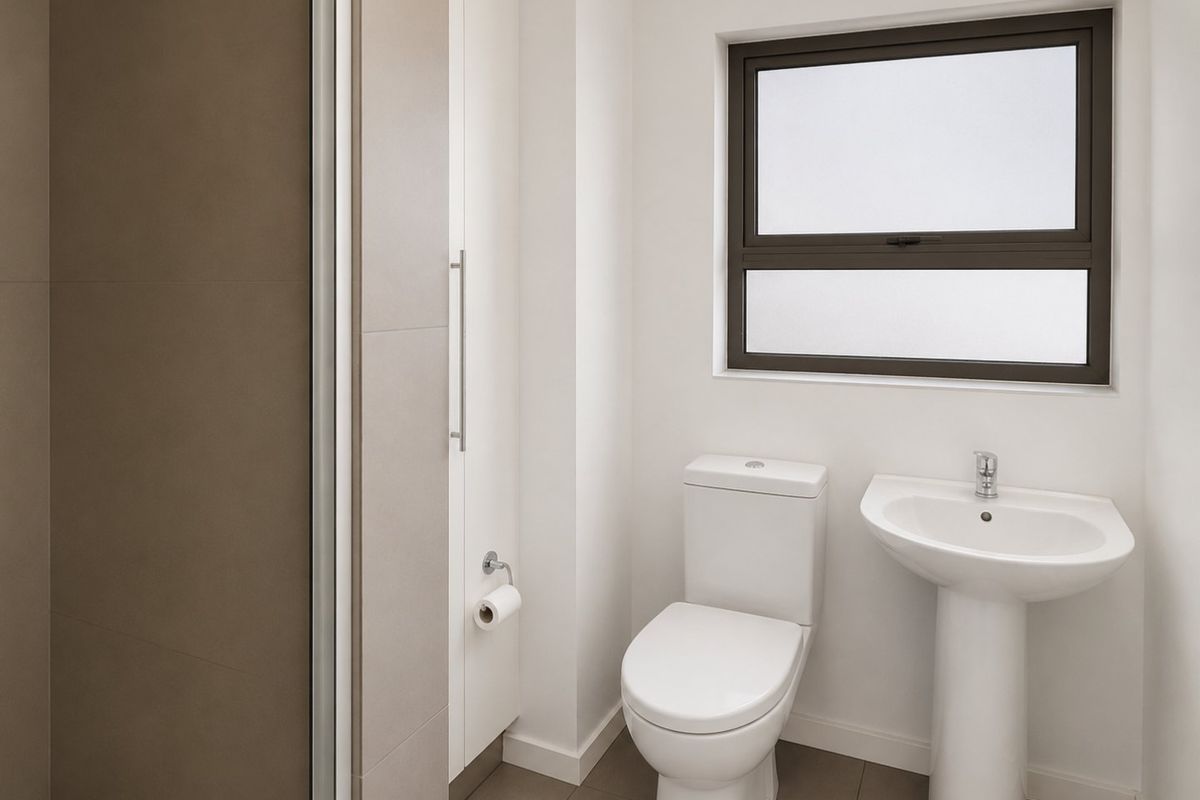 A clean, modern bathroom featuring a pedestal basin, toilet, and shower enclosure with neutral tiling and a frosted window for privacy.