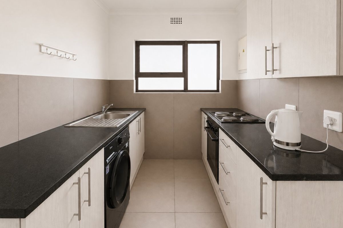 A modern galley-style kitchen featuring light wood cabinetry, dark stone countertops, integrated oven and hob, and neutral tiled finishes.