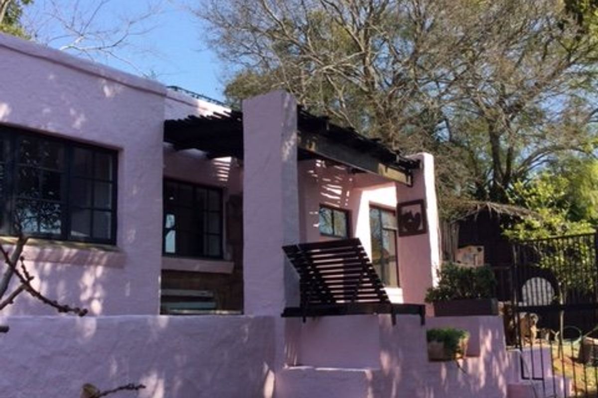 Rear exterior view of a property featuring a pink-painted facade, a pergola-covered patio area, and tiered retaining walls with stone steps.